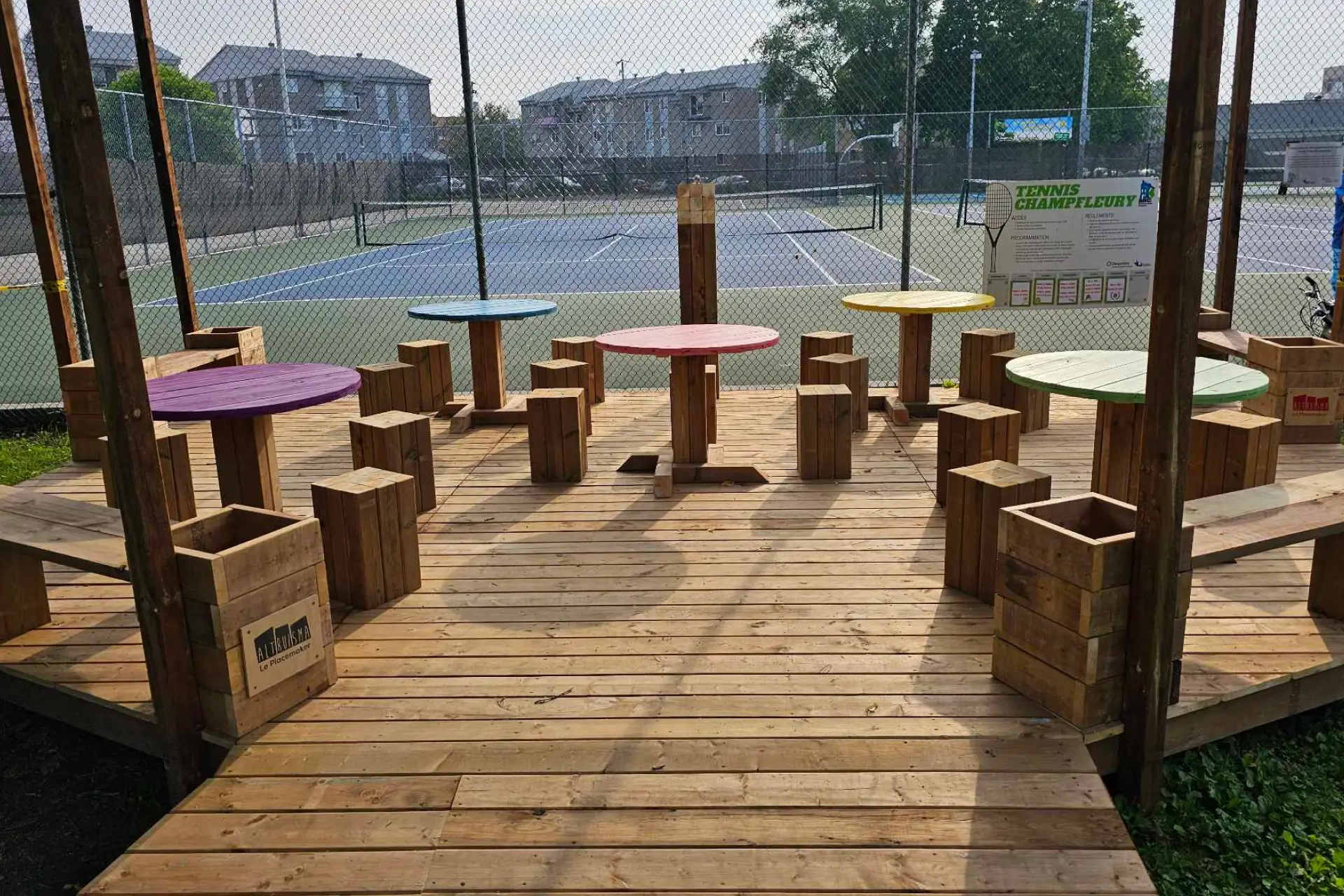 Terrasse en bois animée par des tables rondes colorées près d'un terrain sportif