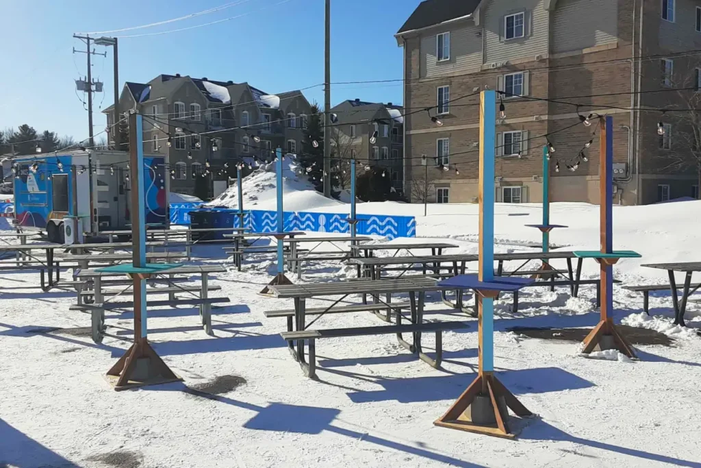 Zone alimentaire avec tables-colonnes peintes et guirlandes lumineuses sur neige au Carnaval d’hiver La Bordée.
