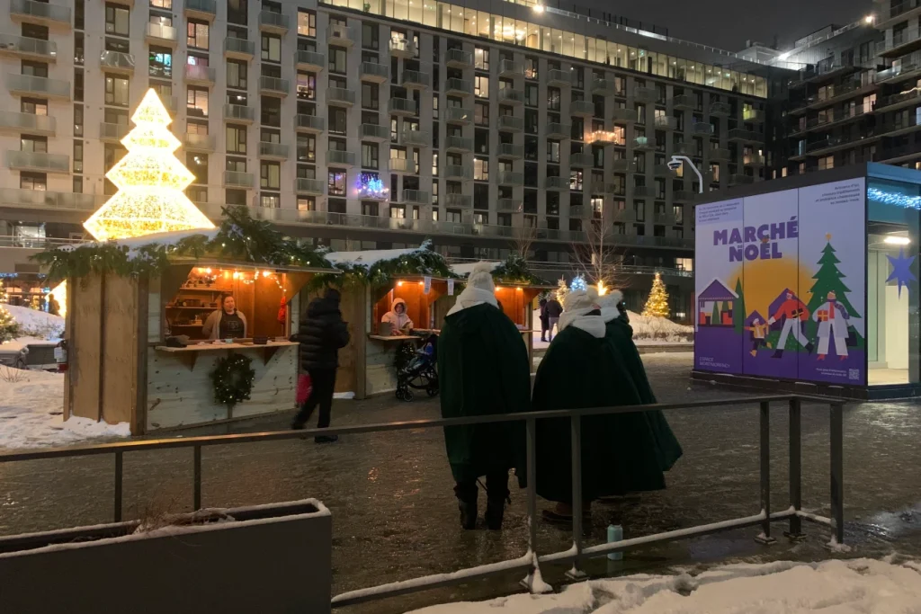 Scène de nuit : grand arbre lumineux et rangée de maisonnettes au Marché de Noël.